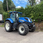 Blue New Holland tractor parked outdoors beside a tall hedge and a small building with a yellow 'SELLS' sign in view.