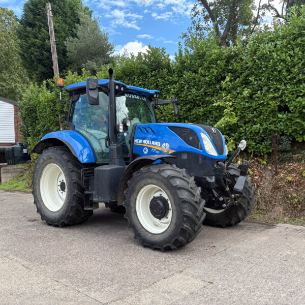 Blue New Holland tractor parked outdoors beside a tall hedge and a small building with a yellow 'SELLS' sign in view.