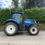 Blue New Holland tractor parked on a paved road with a tall green hedge and trees behind.