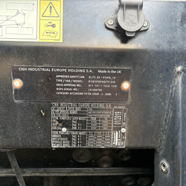 Close-up of a rugged industrial machine surface with multiple metal nameplates, warnings, and a latch; labels include CNH Industrial Europe Holding S.A. and 'Made in the UK' with various model/approval numbers visible.