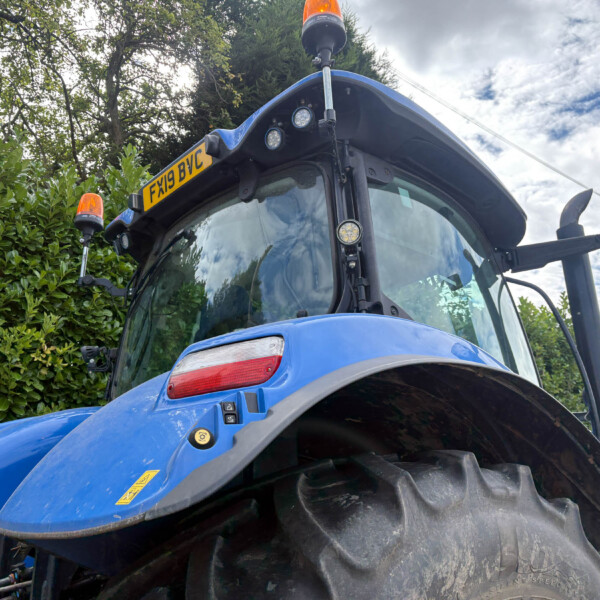 Front view of a blue agricultural tractor with amber beacon lights and license plate FX19 BVC, against green hedges and a cloudy sky.