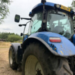 Close-up of a blue tractor front with large muddy tires, side mirror, and orange beacon on top, in a rural field with trees in the background.