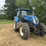 Blue New Holland tractor parked on a dry grassy field with trees in the background, front-right angle.