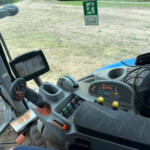 Inside the operator cab of a tractor with the steering wheel, control panel, and joystick visible, looking out at a grassy field.
