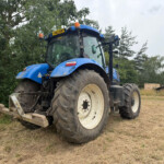 Blue tractor with large tires parked on a dry grassy field beside trees in a rural setting.Front-left view showing orange warning lights on top and dusty wheels.Tractor in a field with dirt path and greenery in the background.Large blue agricultural tractor with mud-covered tires near a wooded area.Blue tractor facing left on dry grass, with trees and pipes visible in the distance.