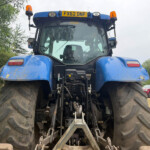 Front view of a large blue agricultural tractor with oversized muddy rear tires, cab in center, and an orange light on the roof; license plate FX62 DNF visible above the windshield.