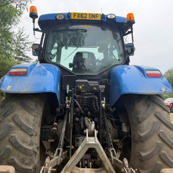 Front view of a large blue agricultural tractor with oversized muddy rear tires, cab in center, and an orange light on the roof; license plate FX62 DNF visible above the windshield.