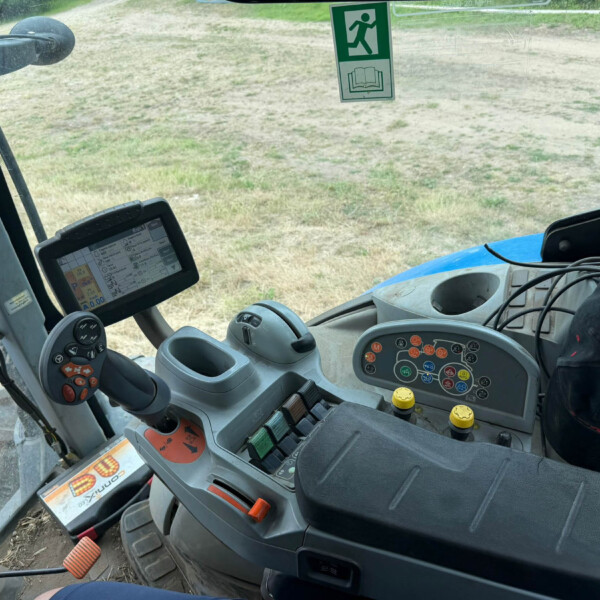 Inside the operator cab of a tractor with the steering wheel, control panel, and joystick visible, looking out at a grassy field.