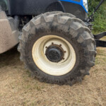 Close-up of a large off-road tractor wheel and tire, with blue New Holland bodywork and dry grass beneath.