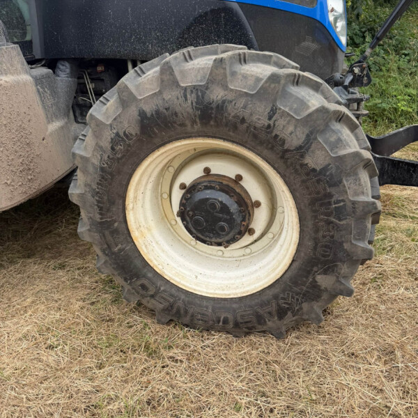 Close-up of a large off-road tractor wheel and tire, with blue New Holland bodywork and dry grass beneath.