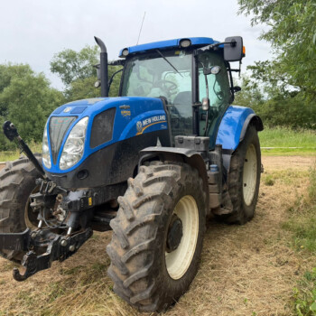 Blue New Holland tractor parked on a dirt field with trees in the background under a cloudy sky in a rural setting.
