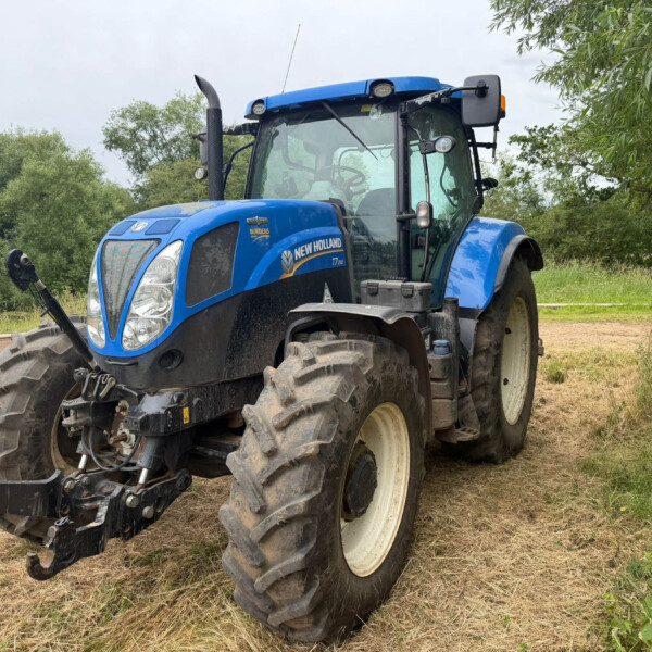 Blue New Holland tractor parked on a dirt field with trees in the background under a cloudy sky in a rural setting.