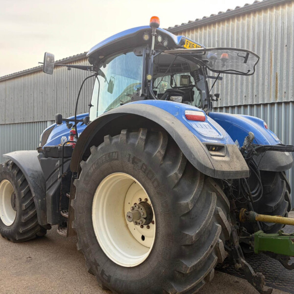 Blue agricultural tractor viewed from the front, parked beside a corrugated metal building with an orange beacon on top and large off-road tires.