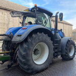 Blue agricultural tractor with large rear tires parked on a grated metal surface, barn-style buildings in the background under a blue sky.