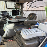 Interior of a farm vehicle cab with a black steering wheel, multiple screens, and a cushioned seat amid dusty controls and dashboards.