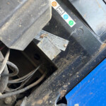 Close-up of a dusty machine frame with warning labels and a metal data plate, showing safety stickers and dirt-streaked surfaces.