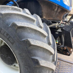 Close-up of a muddy, large tractor rear tire and hitch on a blue tractor × showing hydraulic components and metal linkage.