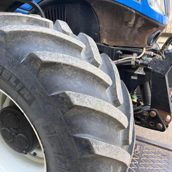 Close-up of a muddy, large tractor rear tire and hitch on a blue tractor × showing hydraulic components and metal linkage.