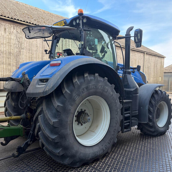 Blue agricultural tractor with large rear tires parked on a grated metal surface, barn-style buildings in the background under a blue sky.