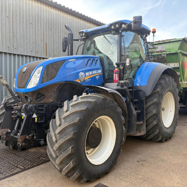 Blue New Holland tractor with large white-walled tires parked beside a corrugated metal wall, attached to a green trailer in a farmyard.