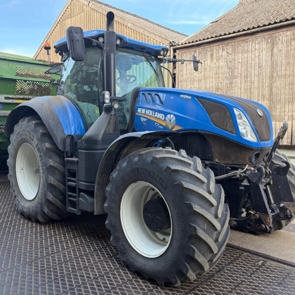 Blue New Holland tractor parked beside a green trailer in a rural yard with wooden barns behind.