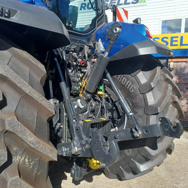 Close-up view of a blue tractor cockpit with exposed hydraulic controls and oversized tires nearby outside a building.