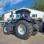 Blue New Holland tractor with oversized tires parked outside a dealership building with a New Holland Agriculture sign.