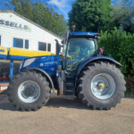 Blue New Holland tractor with oversized tires parked in front of a dealership building labeled 'Russells'. Trees and clear blue sky in background.