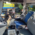Inside a modern truck cab with a gray dashboard, steering wheel, and blue-seat upholstery; a yellow-blue exterior sign is visible through the window.