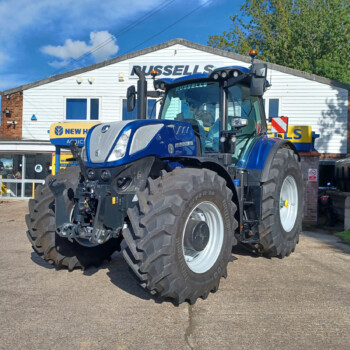 Blue New Holland tractor with oversized tires parked outside an agricultural service shop building with blue doors and signage behind it.
