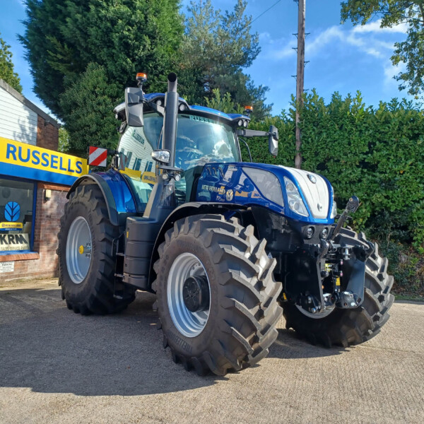 Blue New Holland tractor with large front tires parked outside a building with a yellow and blue Russell's sign in a sunny lot.