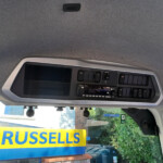 Interior of a vehicle cockpit with a control panel and sun visor, viewed from the passenger seat, through the windshield showing a bright blue-and-yellow 'RUSSELLS' sign outside.
