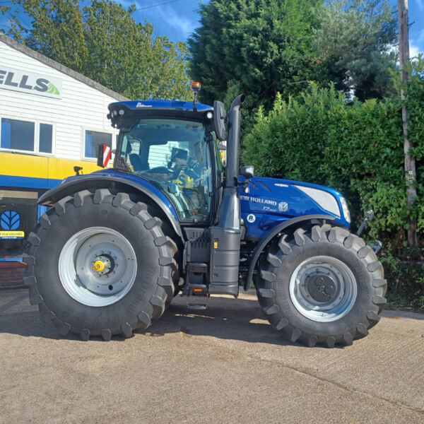 Blue New Holland tractor parked outside a white shop with blue-and-yellow signage; oversized front tires in foreground and green foliage in back