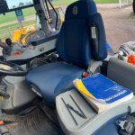 Interior of a blue tractor cab with a control panel, joystick, and a blue cushioned seat; a worn manual sits on the side console; clothes piled on a nearby seat and a JCB machine visible outside.