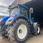 Blue agricultural tractor with large rear tires parked next to a wooden shed, orange beacon lights on top, facing left.