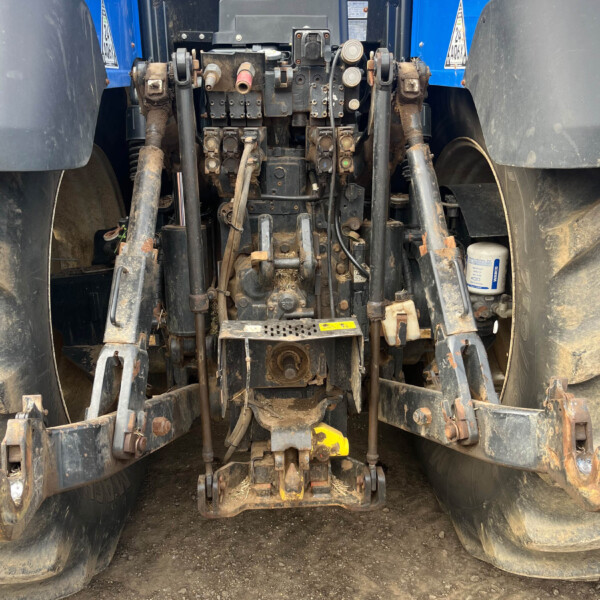 Rear view of a blue tractor showing hydraulic lift arms, hoses, and the three-point hitch on a dirt ground.