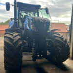 Front view of a large blue tractor inside a shed, with oversized tires and a glass cab, facing the camera. Outside visible are red-tiled rooftops and a cloudy sky.