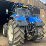 Blue tractor with large muddy tires exiting a dark shed, orange warning lights on top and a yellow license plate visible.
