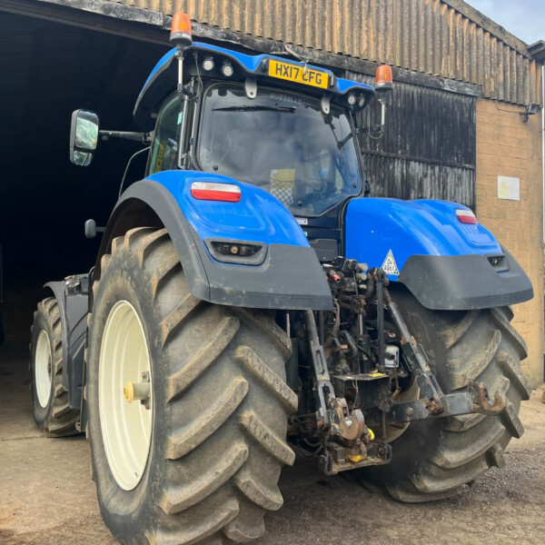 Blue tractor with large muddy tires exiting a dark shed, orange warning lights on top and a yellow license plate visible.