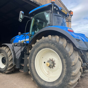 Blue New Holland tractor parked at the entrance of a dark barn or trailer, large rear tires dominating the foreground, orange warning lights on top, rural field in the background.