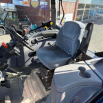 Inside the operator cab of a tractor, with steering wheel, pedals, and control levers, blue seat upholstery visible outside a building site.