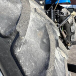 Close-up of a large, dusty tractor tire tread with a metal wheel rim visible on the left and tractor machinery in the background