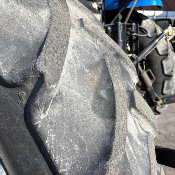 Close-up of a large, dusty tractor tire tread with a metal wheel rim visible on the left and tractor machinery in the background