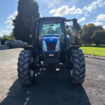 Front view of a blue tractor with large tires parked in a lot, under a blue sky with white clouds and trees in the background.