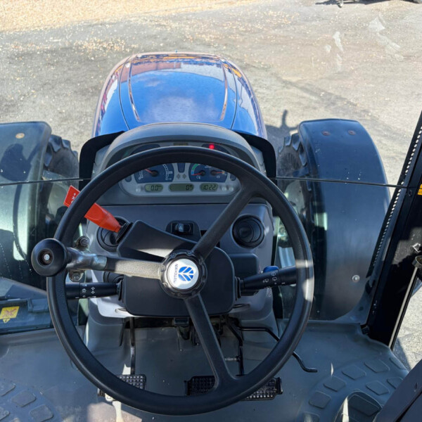 Inside a tractor cab, view of the steering wheel and dashboard with the blue exterior hood visible through the windshield.