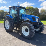 Blue New Holland tractor parked on a paved lot with trees and a bright blue sky behind it.