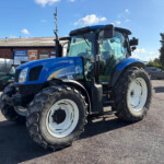 Blue New Holland tractor with oversized front tires parked in a lot, brick building and signs in the background.