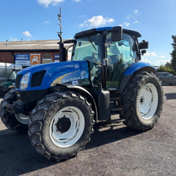 Blue New Holland tractor with oversized front tires parked in a lot, brick building and signs in the background.