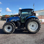 Blue New Holland tractor in a dealership lot with farm equipment and a brick building under a sunny sky.