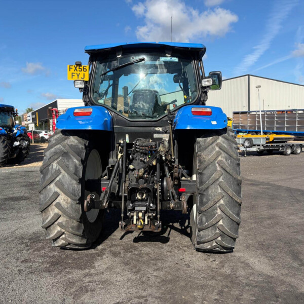 Rear view of a large blue tractor with oversized tires in a dealership lot, other equipment and a white warehouse in the background.